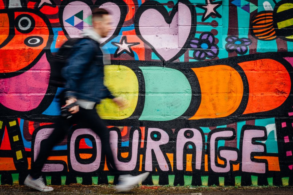Photo of a man walking infront of brightly colored graffiti mural that says courage.