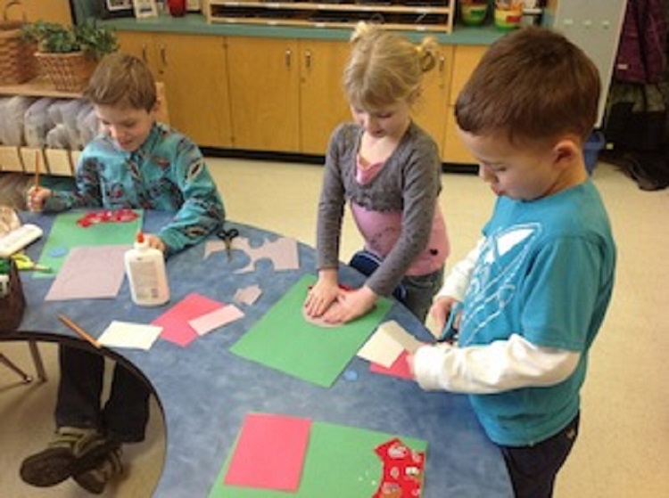 Elementary students standing at table using paper and pencils.