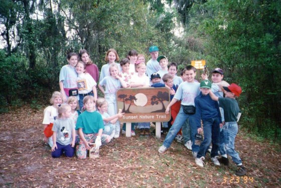 The kids I grew up with - Friendship Bridge Academy. My mom ran a school for homeschooled kids.  Their parents taught some stuff at home, and the rest of our education was like captured in this picture. One room schoolhouse-style, passion-driven, curiosity-based learning where we learned science, history, art, music, and movement by being in nature, acting, singing, dissecting roadkill, making art, playing games, and self-study. Here we are at one of our favorite nature walks, Grassy Pond. I'm in the back center behind the sign, with the bangs." ~ Amber